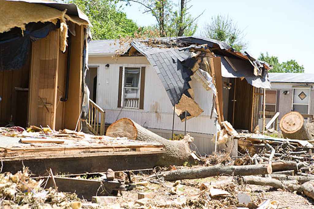 A mobile home with severe roof and wall damage, surrounded by fallen trees and debris, likely caused by a recent storm.