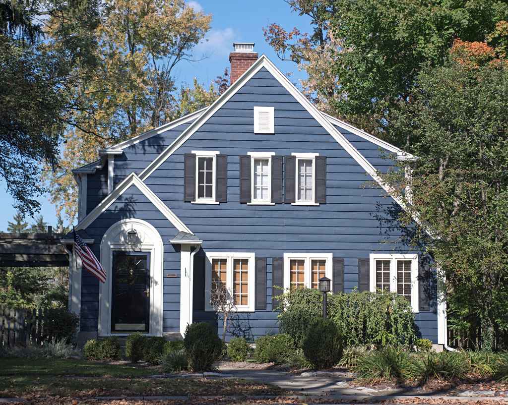 A blue two-story house with white trim, black shutters, and an American flag near the front door, surrounded by trees and shrubs on a sunny day.