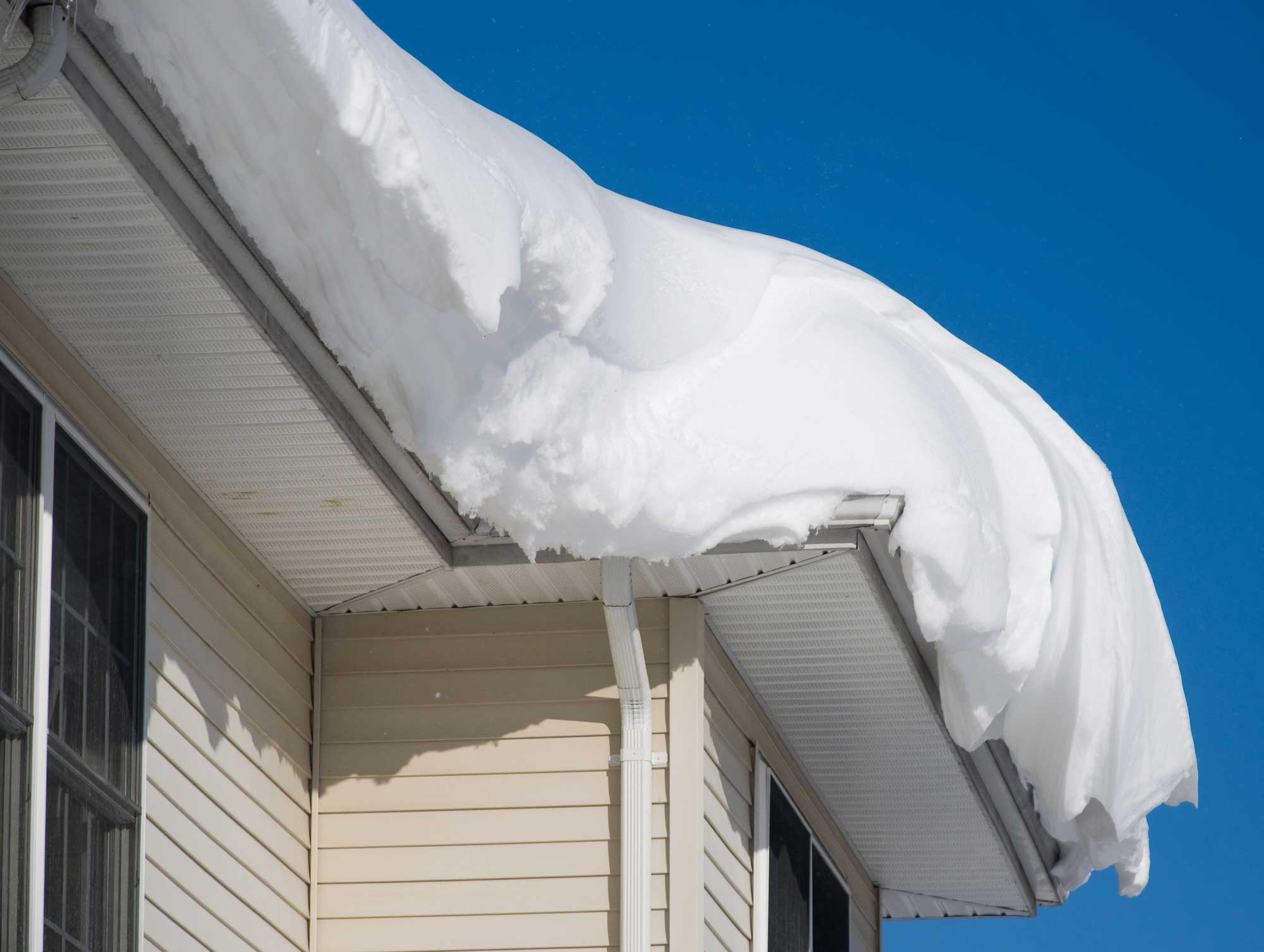 Large accumulation of snow covers the roof and overhang of a beige house against a clear blue sky. Large accumulation of snow covers the roof and overhang of a beige house against a clear blue sky.
