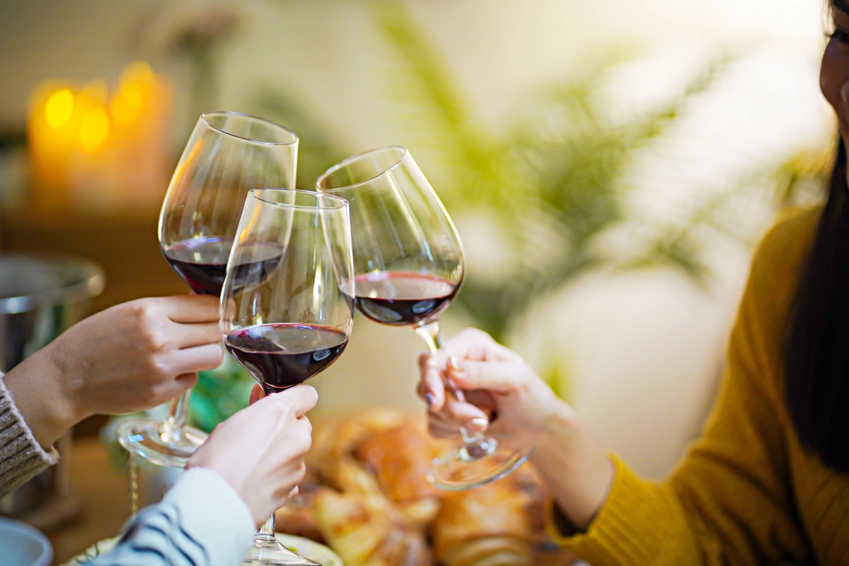 Three people clink wine glasses filled with red wine over a table with bread and pastries, with blurred candlelight and plants in the background.