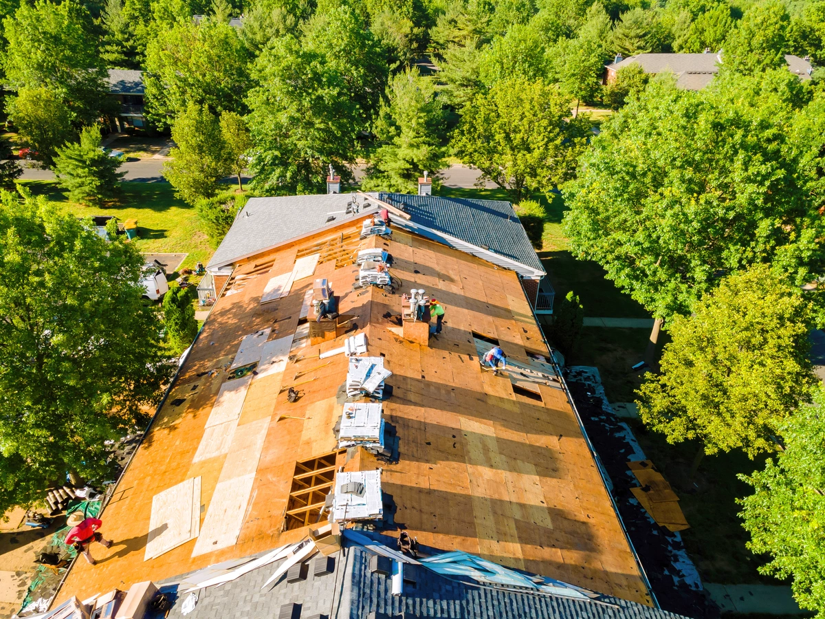Aerial view of workers repairing or replacing shingles on a residential roof surrounded by trees in a suburban neighborhood.