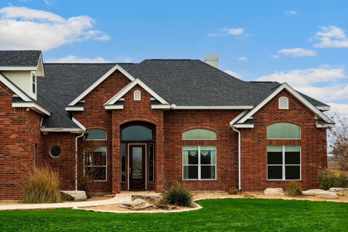 Single-story brick house with black shingle roof, large arched windows, and a dark front door, surrounded by a green lawn and landscaped plants under a partly cloudy sky.