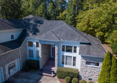 Aerial view of a two-story house with stone and siding exterior, multiple windows, a dark shingled roof, and surrounding trees and shrubs.