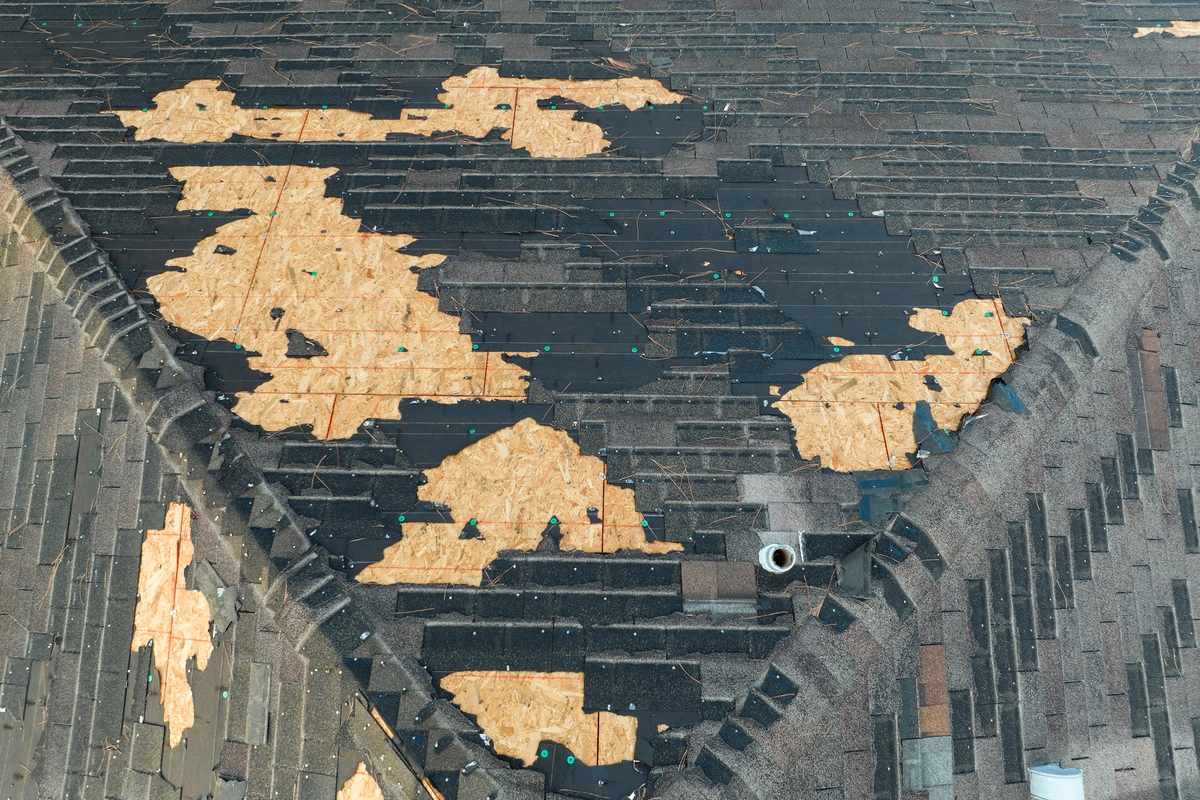 A roof with missing shingles reveals plywood sheathing underneath, exposing sections of the roof to potential weather damage.