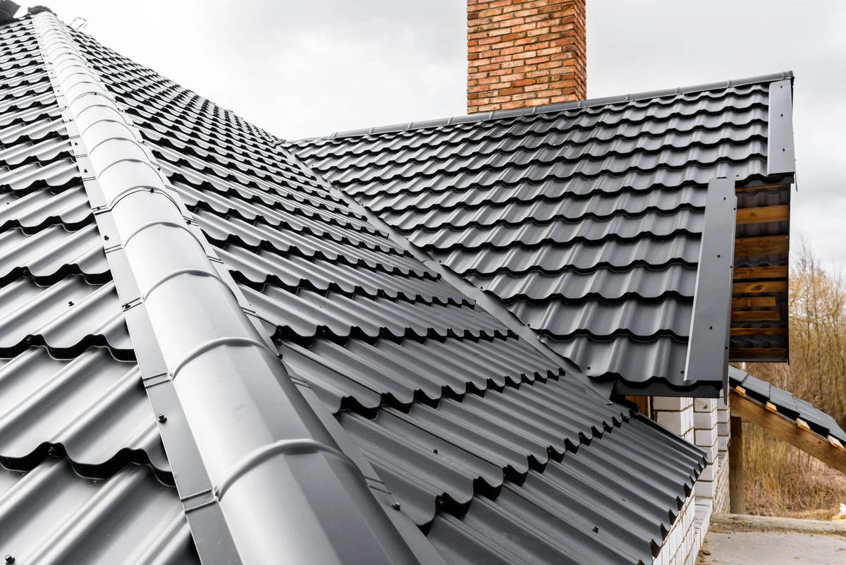 Close-up view of a house roof with black metal roofing sheets and a red brick chimney, under a cloudy sky.