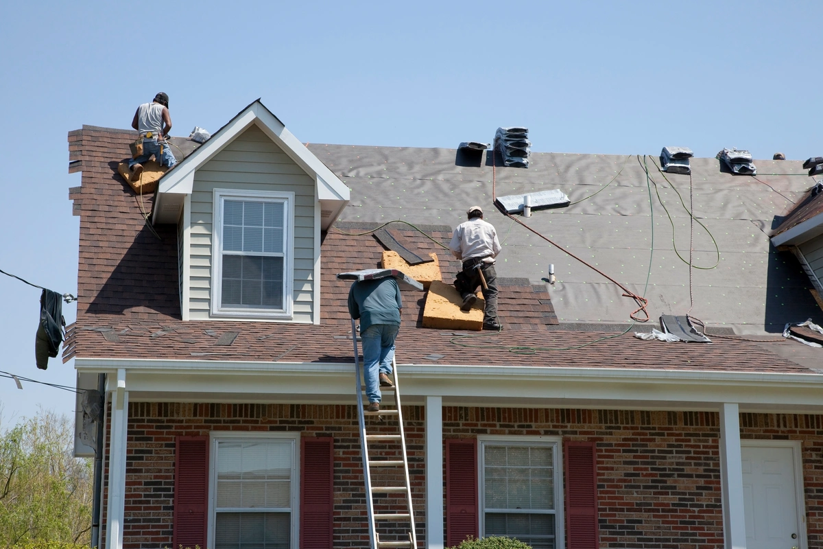 Three workers are installing or repairing shingles on the roof of a brick house, with one worker on a ladder and roofing materials scattered nearby.