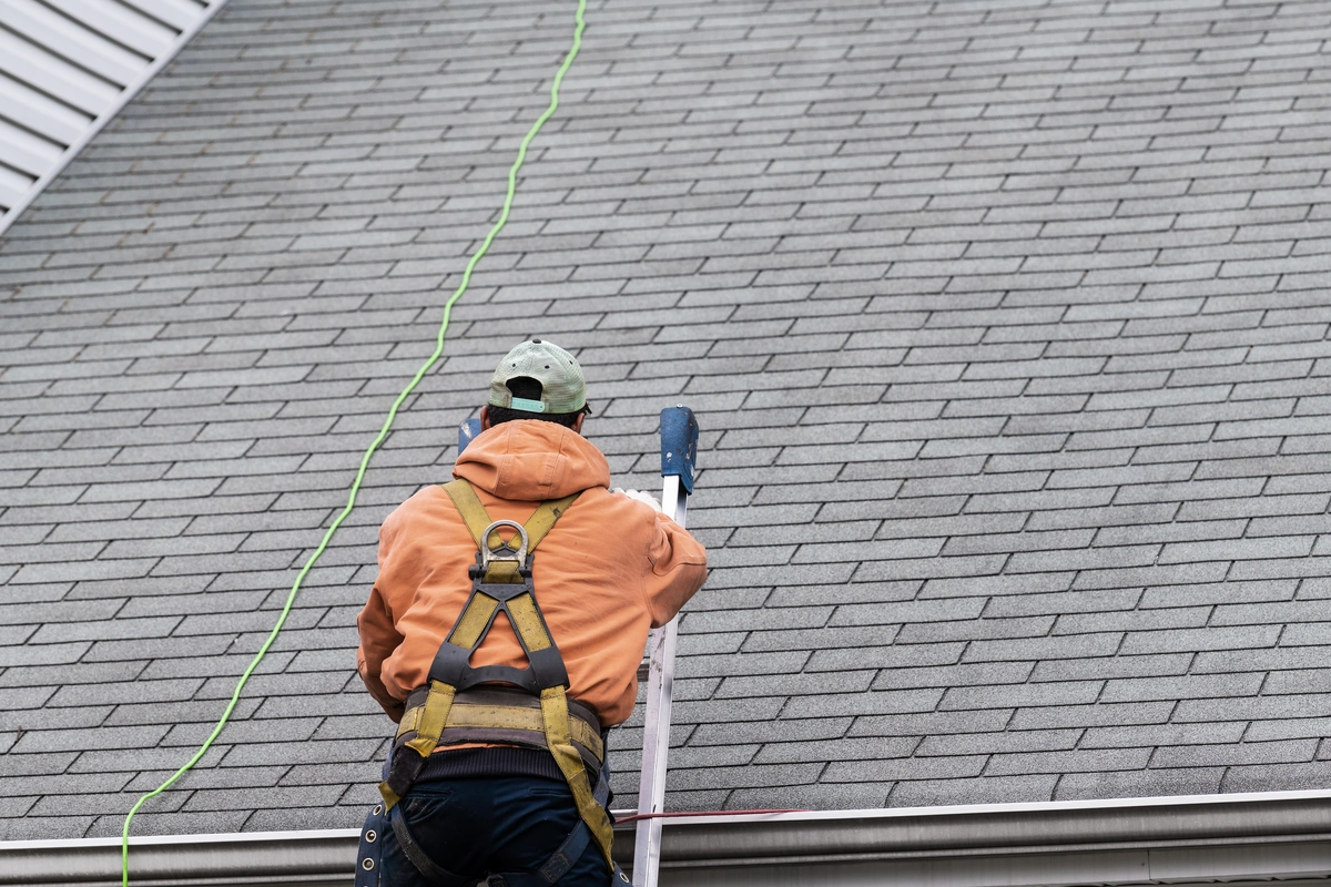 A person in a safety harness climbs a ladder to work on a shingled roof beside a house.