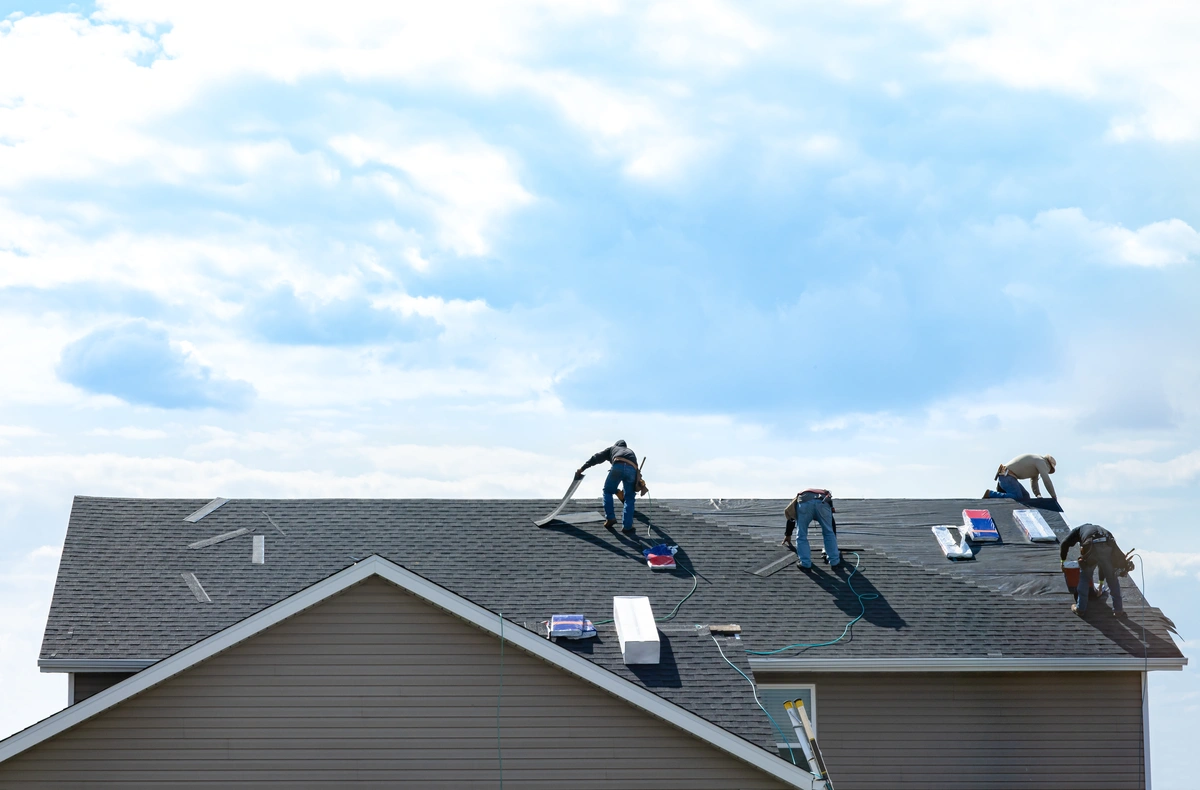 Four workers are installing or repairing shingles on the roof of a two-story house under a bright, partly cloudy sky.