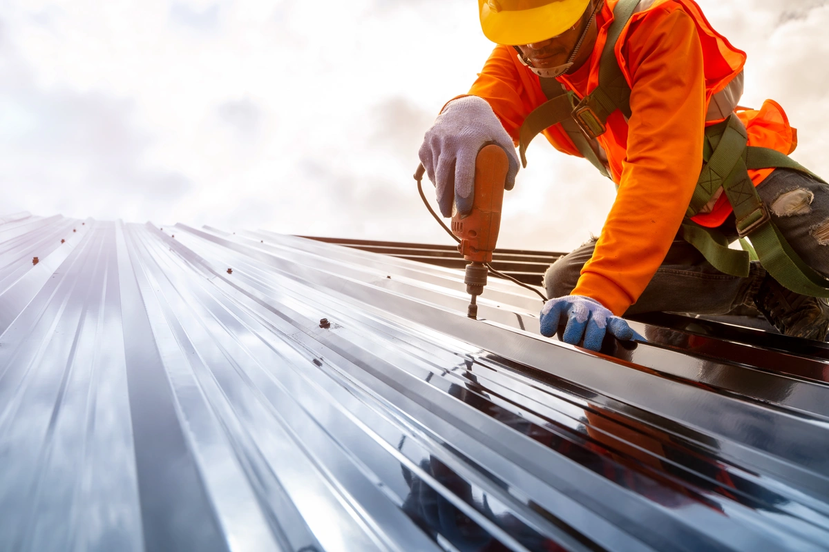 A construction worker in safety gear uses a power drill to fasten screws into a metal roof panel under a cloudy sky.