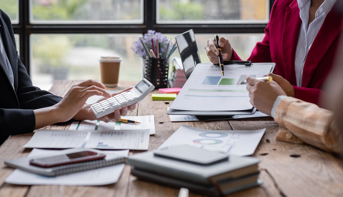Three people sit at a table reviewing financial documents, using a calculator and writing on charts and graphs, with notebooks and coffee on the wooden surface.