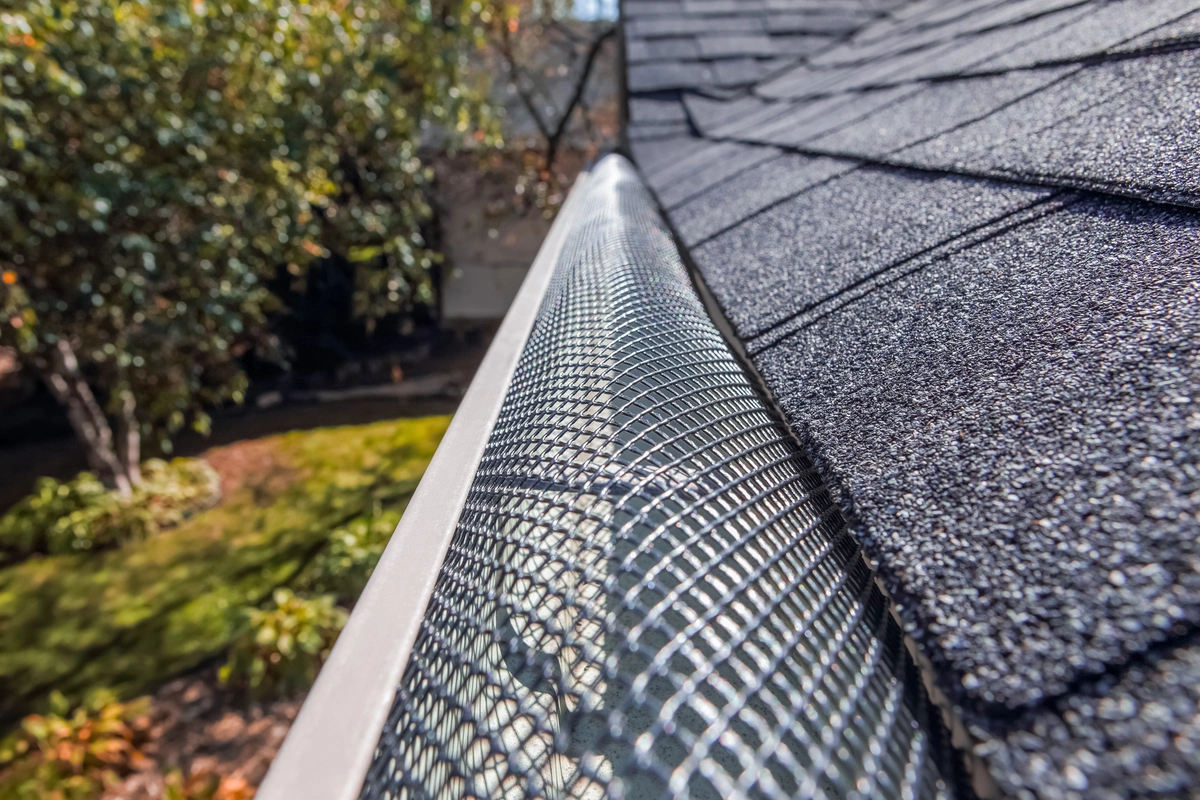 Close-up view of a mesh gutter guard installed along the edge of an asphalt shingle roof, with trees and a landscaped yard in the background.