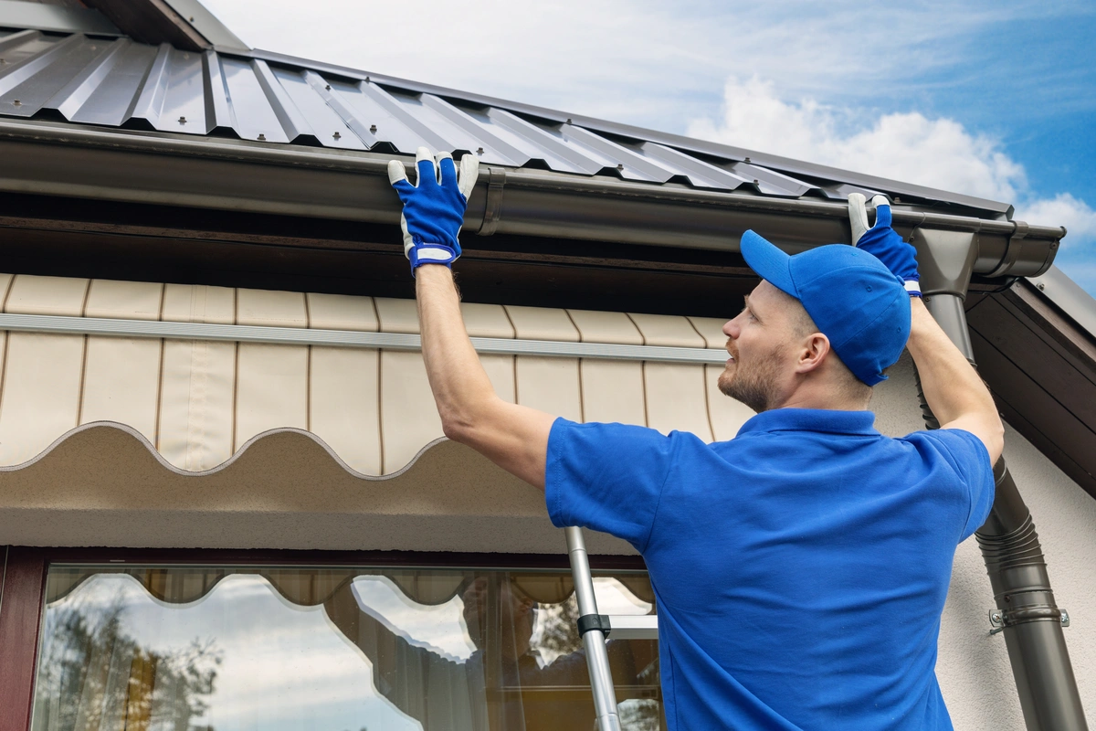 A man in blue work clothes and gloves stands on a ladder, installing or repairing a rain gutter on the roof edge of a house.