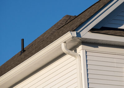 Close-up of a house exterior showing white vinyl siding, a white gutter and downspout, and a gray asphalt shingle roof against a clear blue sky.