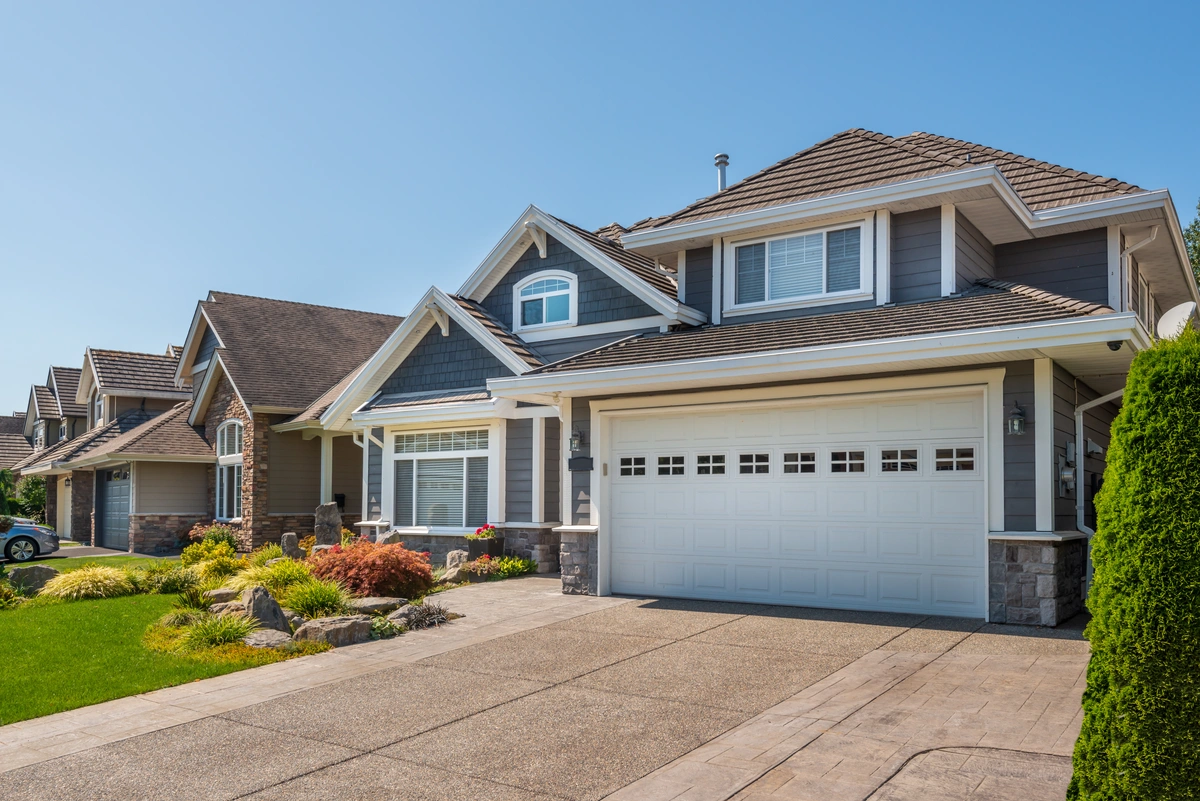 Suburban house with gray siding, white trim, a double garage door, manicured lawn, and landscaping, photographed on a clear, sunny day.