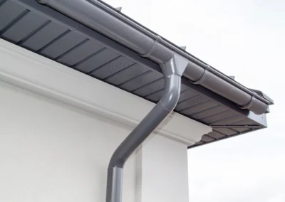 Close-up of a modern house’s corner showing a black rain gutter system attached to the roof and a downspout running along a white exterior wall.