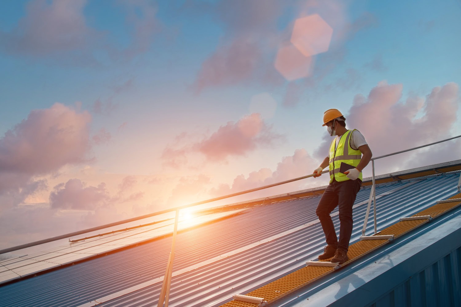 A construction worker in a safety vest and helmet stands on a metal rooftop at sunset, holding a guardrail and looking at the horizon.