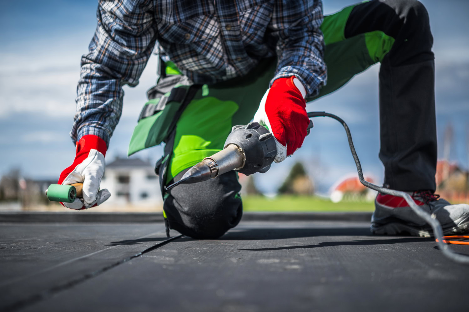 Worker in protective gloves kneeling on a flat roof, using a heat welding tool to seal roofing membrane seams.