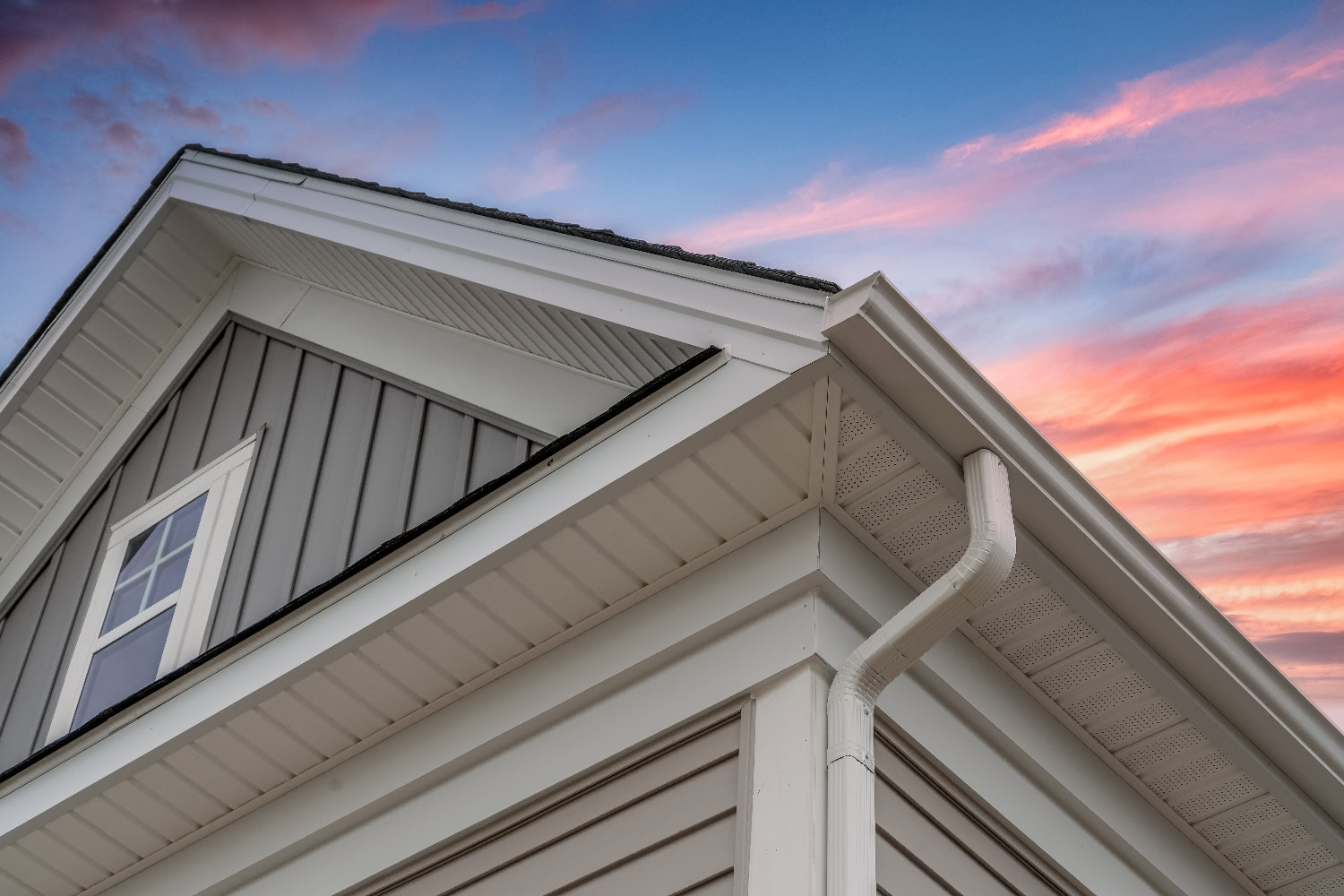 Close-up of the edge of a house with white siding and gutters, featuring a window and gable roof, set against a colorful sunset sky.
