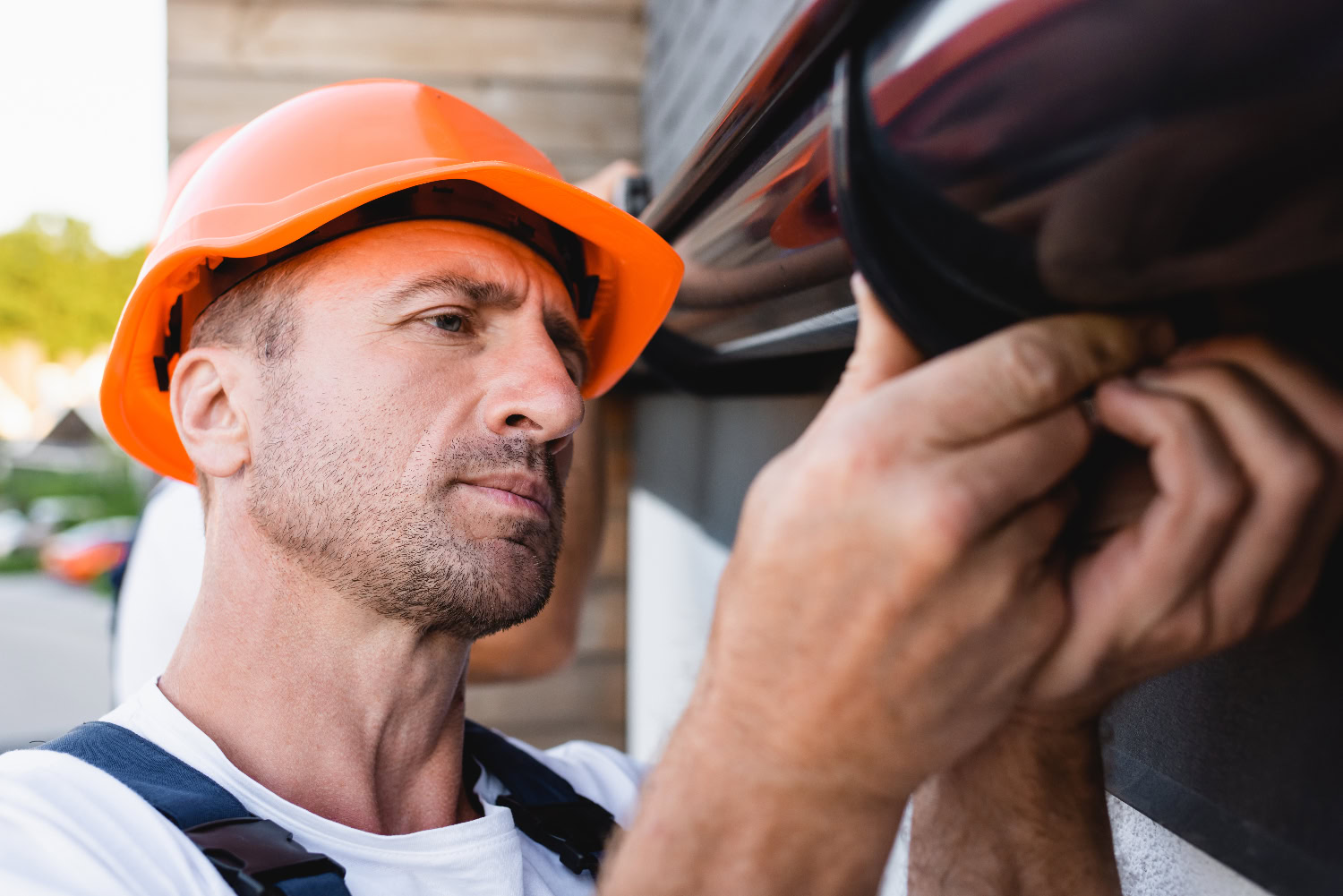 Man wearing an orange hard hat and white shirt installs or inspects a black fixture on an exterior wall, focusing intently on his work.