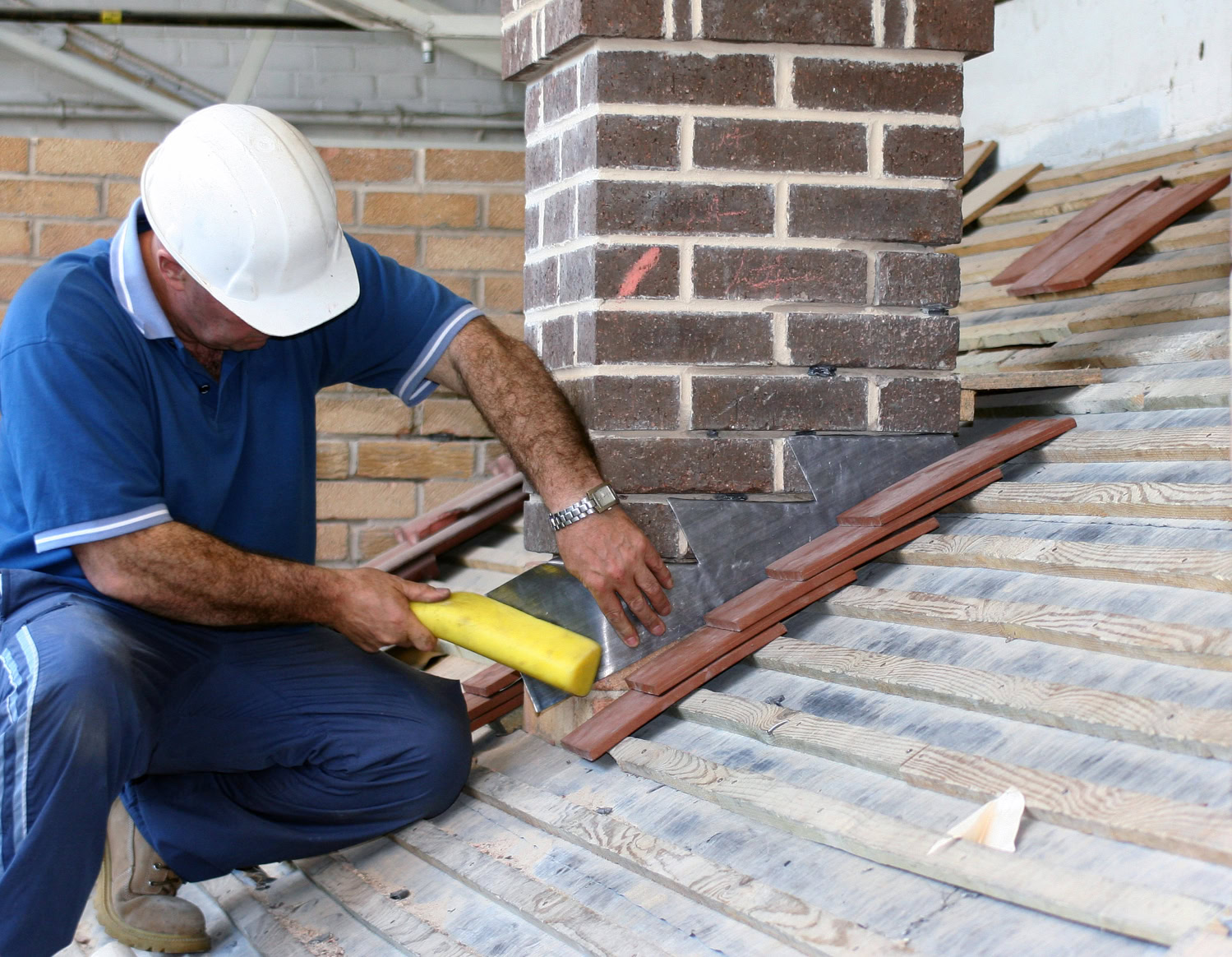 A construction worker in a white hard hat installs roof tiles around a brick chimney on a sloped wooden roof.