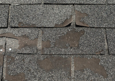 Close-up of weathered asphalt roof shingles with significant granule loss, curling, and visible damage on several shingles.