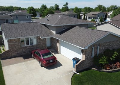 A single-story suburban house with brick and siding exterior, an attached two-car garage, and a red car parked in the driveway. Green lawns and neighboring houses are visible.
