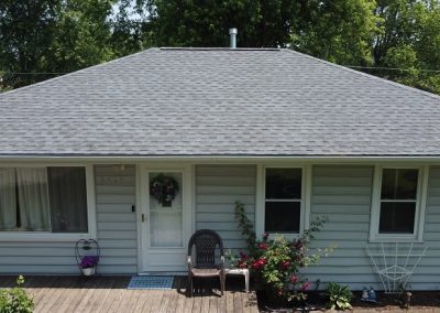 Single-story house with gray siding and a gray shingled roof, featuring a small porch, two chairs, potted plants, and a doormat near the white front door.