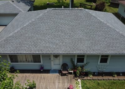 Aerial view of a single-story house with a gray shingled hip roof, front porch, two chairs, flowers, and a detached garage in the background.