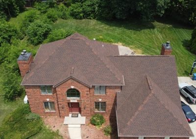 Aerial view of a two-story brick house with a brown roof, surrounded by grass and trees. Several cars are parked in the driveway.