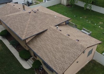 Aerial view of a tan house with a brown shingled roof, surrounded by a white fence and green lawn, with nearby driveways and neighboring houses.