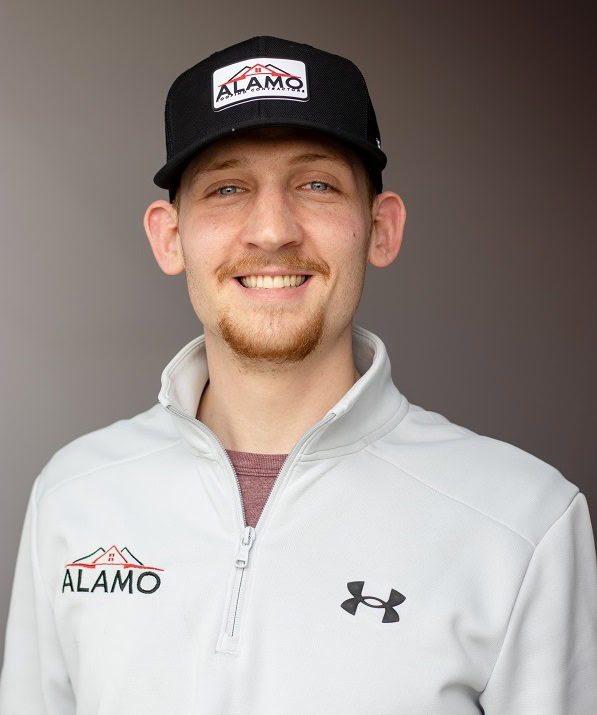 A man wearing a black Alamo cap and a light gray Under Armour jacket stands smiling in front of a neutral background.