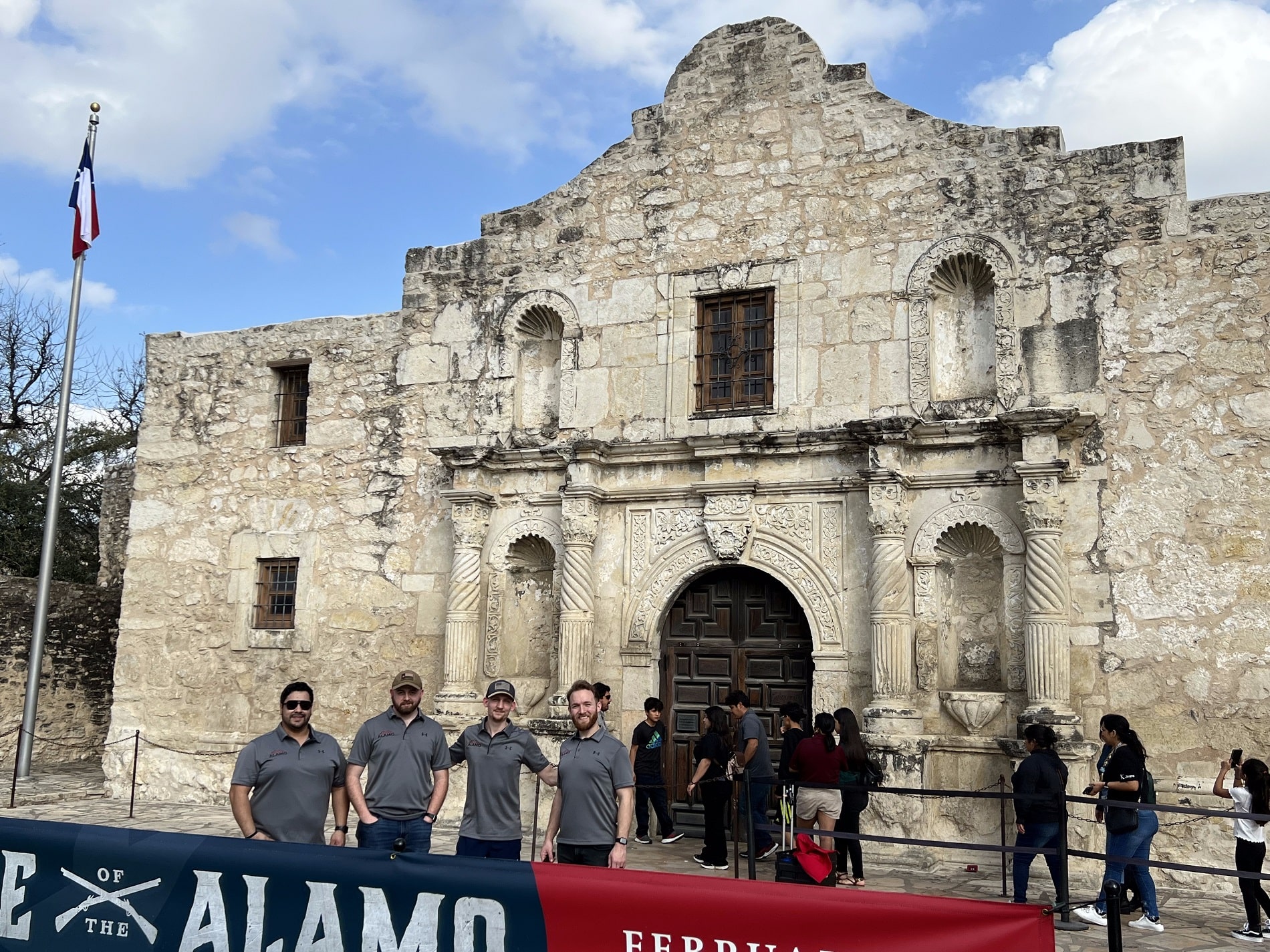 Four men stand in front of the historic Alamo building in San Antonio, Texas, with other visitors nearby and a Texas flag on the left.